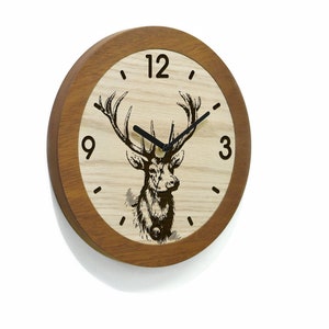 Puede incluir: Un reloj de pared de madera con un marco de madera marrón y una cara de madera clara. La esfera del reloj presenta una ilustración en blanco y negro de un ciervo con cuernos. La esfera del reloj también tiene números negros para 12, 3, 9, y las manecillas son negras.