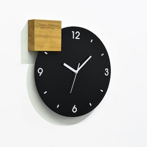 Può includere: Un orologio nero con numeri e lancette bianchi, montato su una parete bianca. Un piccolo blocco di legno con il testo "Oliver + Melissa" è attaccato all'orologio.