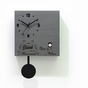 May include: A gray wooden cuckoo clock with a black pendulum and a white cuckoo bird. The clock face has black Roman numerals and hands. The clock is engraved with the text "Eat Drink and be Married" and the names "Oliver & Melissa".