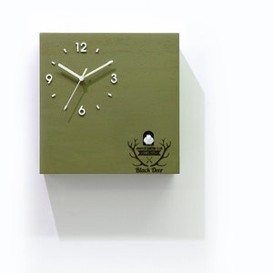 May include: A green wooden cuckoo clock with a white face and black hands. The clock has a black deer logo with the text "Black Deer" below it. The clock is mounted on a white wall.