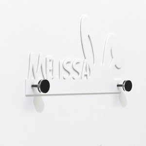 以下が含まれることがあります： 名前「メリッサ」がくり抜かれた白い木製の壁掛けフックと、黒いノブが2つ。フックはウサギの耳の形をしています。