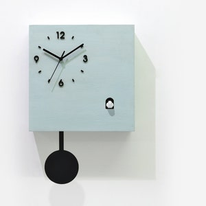 May include: A light blue square cuckoo clock with black hands and a black pendulum. The clock face has black Roman numerals and a small white bird cutout. The clock is labeled "Cuckoo Clocks Time Box by Sol Pixie Dust".