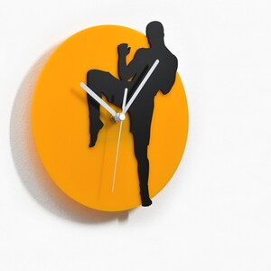 May include: A wall clock featuring a black silhouette of a martial artist in a kicking pose against a bright orange circular background. The clock hands are white. The clock is mounted on a white wall.