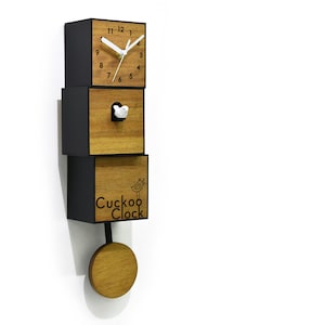 Könnte beinhalten: Eine Holzkuckucksuhr mit schwarzem Zifferblatt und einem weißen Vogel. Die Uhr besteht aus drei übereinander gestapelten Holzblöcken. Der untere Block trägt die Worte "Cuckoo Clock" eingraviert. Der mittlere Block hat ein kleines Vogelloch mit einem weißen Vogel, der herausguckt. Der obere Block hat ein Uhrwerk mit schwarzen Zahlen und Zeigern. Die Uhr hat ein Pendel, das hin und her schwingt.