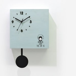 May include: A light blue cuckoo clock with a black pendulum and a white cat illustration. The clock face has black hands and black numbers. The clock reads "Home is where the cat is".