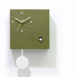 Op de afbeelding: Een groene vierkante koekoeksklok met witte wijzers en cijfers. De klok heeft een wit slinger en een kleine witte vogel die uit het gezicht van de klok springt.