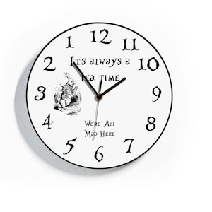 May include: A white round clock with black numbers and hands. The clock face features a black and white illustration of the White Rabbit from Alice in Wonderland. The text on the clock reads "It's always a tea time. We're all mad here."