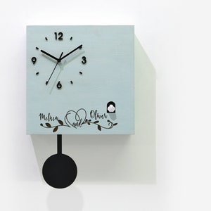 May include: A light blue cuckoo clock with a pendulum and a black clock face. The clock face has black hands and black Roman numeral markings. The clock has a small white bird cutout on the front. The clock is personalized with the names "Melissa and Oliver" and a heart design.