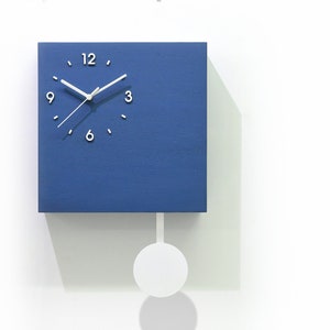 May include: A blue square clock with white hands and numbers. The clock has a white pendulum and is hanging on a white wall. The text "Cuckoo Clocks" and "Time Box" is visible in the image.