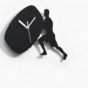 May include: Black silhouette wall clock featuring a person pushing a large tire. The clock face is on the tire and has white hands.