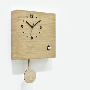 Puede incluir: Un reloj de cuco de madera con un péndulo y un pequeño pájaro que sale. La esfera del reloj tiene números y manecillas negros. El reloj está hecho de madera clara y tiene una forma cuadrada.