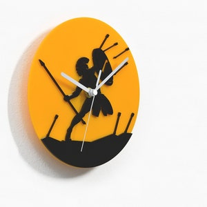 May include: A round wall clock with a yellow background and a black silhouette of a warrior with a spear and shield. The clock hands are white. The clock is mounted on a white wall.