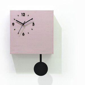 May include: A pink square cuckoo clock with black hands and a black pendulum. The clock face has black Roman numeral markings. The clock is labelled "Cuckoo Clocks Time Box by Sol Pixie Dust".
