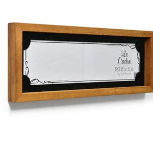 May include: A wooden frame with a black mount and a white paper insert. The paper insert has a decorative border and the text "Le Cadre 20.8 x 5.6 by Sol Studio".