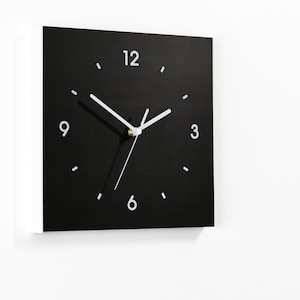 Puede incluir: Un reloj cuadrado negro con números y manecillas blancas. El reloj está colgado en una pared blanca.