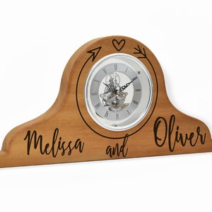 Puede incluir: Reloj de chimenea de madera con una esfera plateada y agujas negras. El reloj está grabado con los nombres "Melissa y Oliver" y una flecha con un corazón.