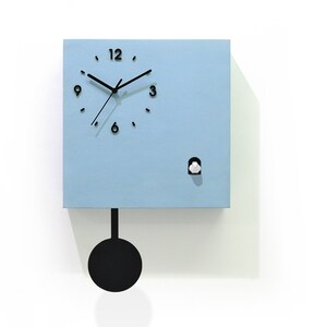 May include: A light blue cuckoo clock with black hands and numbers. The clock has a white bird that pops out of the front. The clock is hanging on a white wall with a black pendulum.