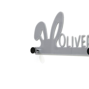May include: Grey bunny-shaped coat rack with the name "Oliver" cut out. The rack has two black knobs and is mounted on a white wall.