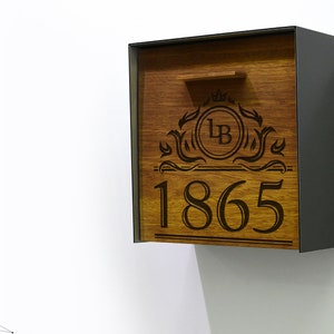 Puede incluir: Un buzón de madera con marco de metal negro. El buzón tiene un diseño decorativo con las iniciales "LB" y el año "1865" grabados.
