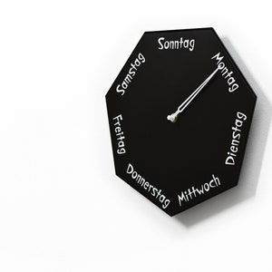 Könnte beinhalten: Eine schwarze achteckige Uhr mit weißer Beschriftung, die die Wochentage auf Deutsch angibt. Die Uhrzeiger zeigen auf Montag.