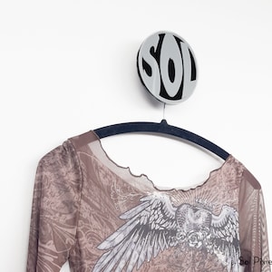 Könnte beinhalten: Ein braunes, langärmliges Netzshirt mit einem weißen Aufdruck eines Engels mit Flügeln und einem Herzen. Das Shirt hängt an einem schwarzen Kleiderbügel mit einem runden, silbernen Schild, auf dem in schwarzen Buchstaben "SOLD" steht.