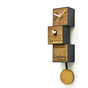 May include: A wooden cuckoo clock with a black frame. The clock face has black Roman numerals and black hands. The clock has a small white bird that pops out of the top of the clock. The clock is labeled "Cuckoo Clock" and has a pendulum.