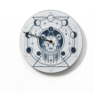 May include: A white circular clock with a black bear face in the center. The bear is surrounded by a geometric design with moons and stars. The clock hands are black.