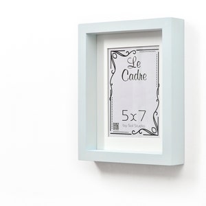 May include: A light blue picture frame with a white mount and a 5x7 inch opening. The frame has a decorative border and the text "Le Cadre by Sol Studio" is printed on the mount.