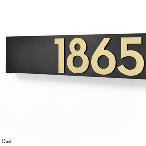 Könnte beinhalten: Ein schwarzes rechteckiges Schild mit der Nummer 1865 in goldenen, erhabenen Buchstaben.