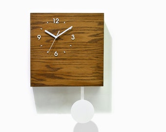Puede incluir: Un reloj cuadrado de madera con una esfera blanca y agujas blancas. El reloj tiene un péndulo y está colgado en una pared blanca. El reloj está hecho por Time Box Cuckoo Clocks.