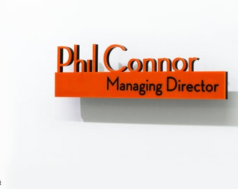 Könnte beinhalten: Ein orangefarbenes und schwarzes Schild mit dem Text "Phil Connor Managing Director" an einer weißen Wand.