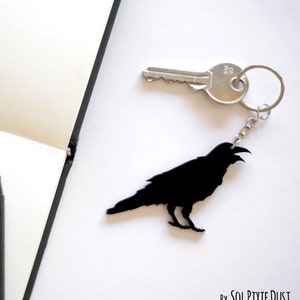May include: Black crow silhouette keychain with a silver key ring and a silver key. The keychain is on a white surface. By Sol Pixie Dust.