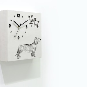 Puede incluir: Reloj cuadrado blanco con agujas negras y números negros. La esfera del reloj tiene una ilustración en blanco y negro de un dachshund y el texto "Love my" con corazones.