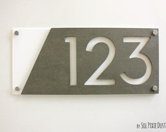 Puede incluir: Un letrero de número de casa moderno con los números 123 recortados en un panel de metal gris. El panel está montado sobre un fondo blanco con un recorte diagonal.