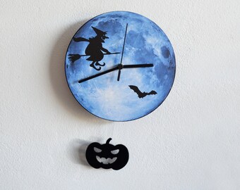 Peut inclure: Horloge murale sur le thème d'Halloween. Elle présente un fond de lune bleue avec une silhouette noire de sorcière sur un balai et une chauve-souris. Une citrouille noire est suspendue en dessous.