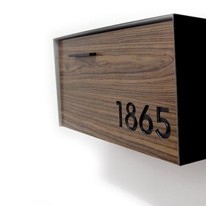 May include: A wooden mailbox with a black metal frame and a black metal handle. The mailbox has a wood grain pattern and the number 1865 is cut out of the wood.