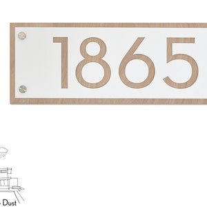 Könnte beinhalten: Ein rechteckiges weißes Schild mit einem Holzrahmen und der Nummer 1865, die in der Mitte ausgeschnitten ist. Das Schild ist mit vier silbernen Schrauben an einer Wand befestigt.