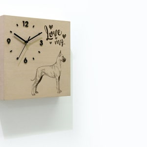 May include: Square wooden wall clock with black hands and numerals. The clock face features a Great Dane dog illustration and the words "Love my". The clock is mounted on a white wall.