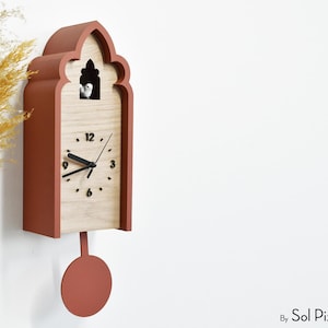 May include: A terracotta-colored cuckoo clock with a light wood face and black clock hands. The clock has a decorative arched top and a circular pendulum. A small bird is visible inside the clock. The clock is mounted on a white wall.