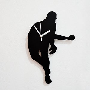 May include: Black silhouette of a baseball player pitching, used as a clock on a white wall. The clock hands are white.