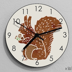 May include: A round white clock with black numbers and hands features a brown squirrel holding a nut. The clock has a white background with a brown squirrel illustration in the center. The text "By SOL PIXIE DUST" is printed at the bottom of the clock.