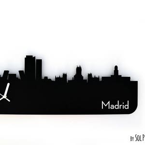 May include: Black and white silhouette of the Madrid skyline with a clock face in the lower left corner. The clock face has white hands and a black background. The text "Madrid" is printed in white below the skyline. The text "By SOL PIXIE DUST" is printed in white in the lower right corner.