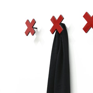 Op de afbeelding: Drie rode haken in de vorm van een X, gemonteerd op een witte muur. Een zwarte sjaal hangt aan een van de haken.