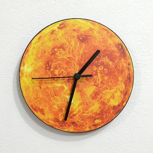 May include: A round clock with a black face and hands. The clock face features a detailed image of the planet Venus in shades of orange and yellow.