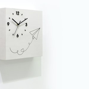 May include: White square clock with black hands and numbers. The clock face has a paper airplane with a heart-shaped tail flying out of it. The clock is mounted on a white wall.