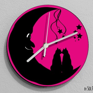 Puede incluir: Un reloj redondo rosa con siluetas negras de dos gatos besándose bajo una luna creciente. Las manecillas del reloj son blancas y hay pequeñas estrellas blancas dispersas alrededor de la esfera del reloj. El texto "By SOL PIXIE DUST" está en la parte inferior del reloj.