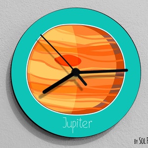 Puede incluir: Un reloj redondo turquesa con una esfera y agujas negras. La esfera presenta una imagen estilizada de Júpiter, el planeta más grande de nuestro sistema solar, con rayas naranjas y marrones. El texto "Jupiter" está impreso debajo de la imagen. El texto "By SOL PIXIE DUST" está impreso debajo del reloj.
