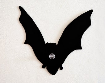 Op de afbeelding: Zwarte silhouet van een vleermuis met een zilveren metalen knoop in het midden. De vleermuis is gemonteerd op een witte muur.