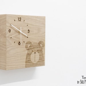 May include: A wooden clock with a bear design. The clock face has black numbers and a white hour and minute hand. The clock is made of light brown wood and has a square shape.