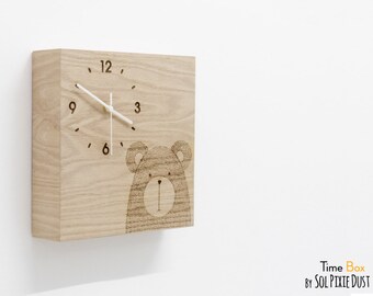 Puede incluir: Un reloj de madera con un diseño de oso. La esfera del reloj tiene números negros y una aguja de hora y minutos blanca. El reloj está hecho de madera marrón claro y tiene una forma cuadrada.